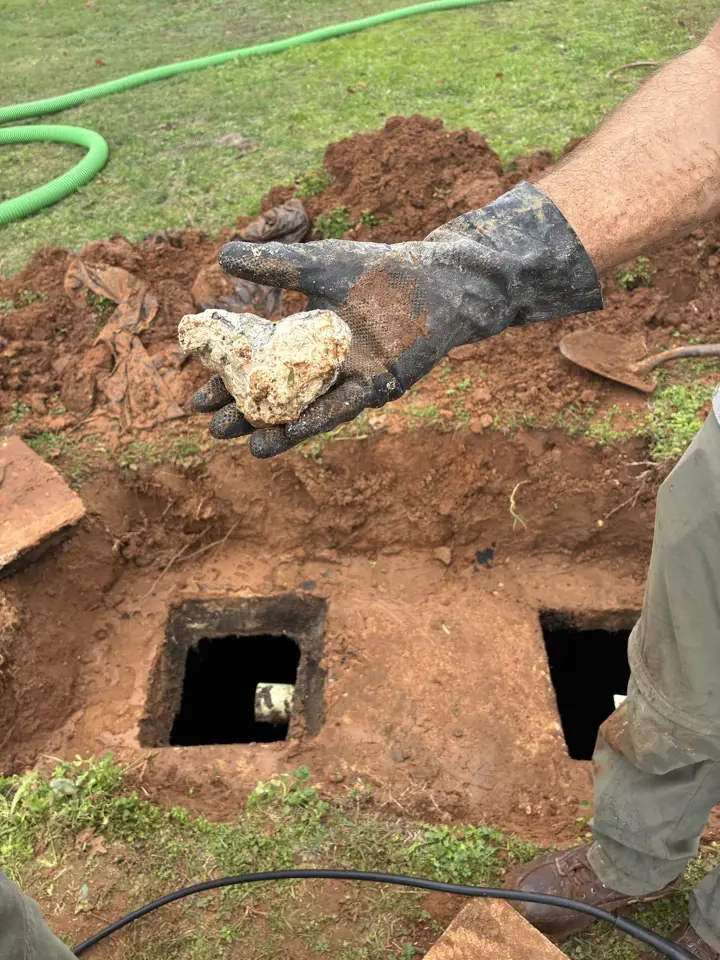 Gloved hand holding a pale chunk of rock dug out beside two open septic tank access holes